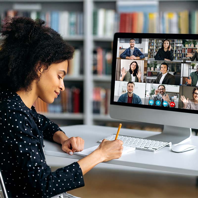 Association header 800x800px V2 Video call, online conference using app and computer. African American female, studying remotely, watches an online lecture, taking notes, multiracial smiling people on the computer screen