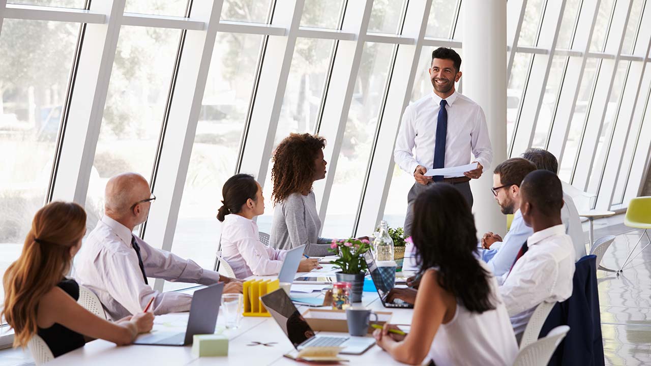 Collaborative leadership 1280x720px young professionla may with suit and tie standing presenting in ftront of a meeting table presenting to a group of work collegues