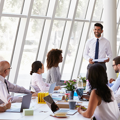 Collaborative leadership 400x400px young professionla may with suit and tie standing presenting in ftront of a meeting table presenting to a group of work collegues