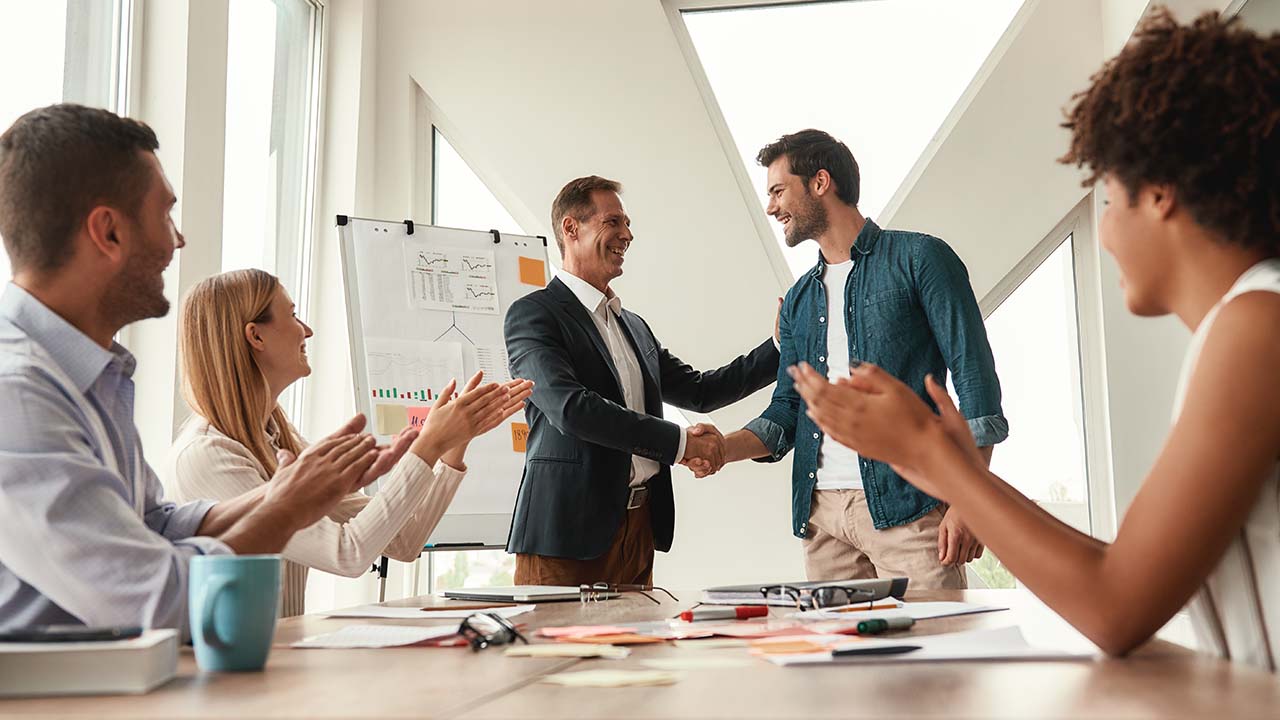 Elevate Capability 1280x720px group of people in meeting room. Male manager shaking hand of employee and congratualting him