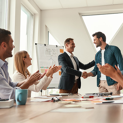 Elevate Capability Mobile 400x400px group of people in meeting room. Male manager shaking hand of employee and congratualting him