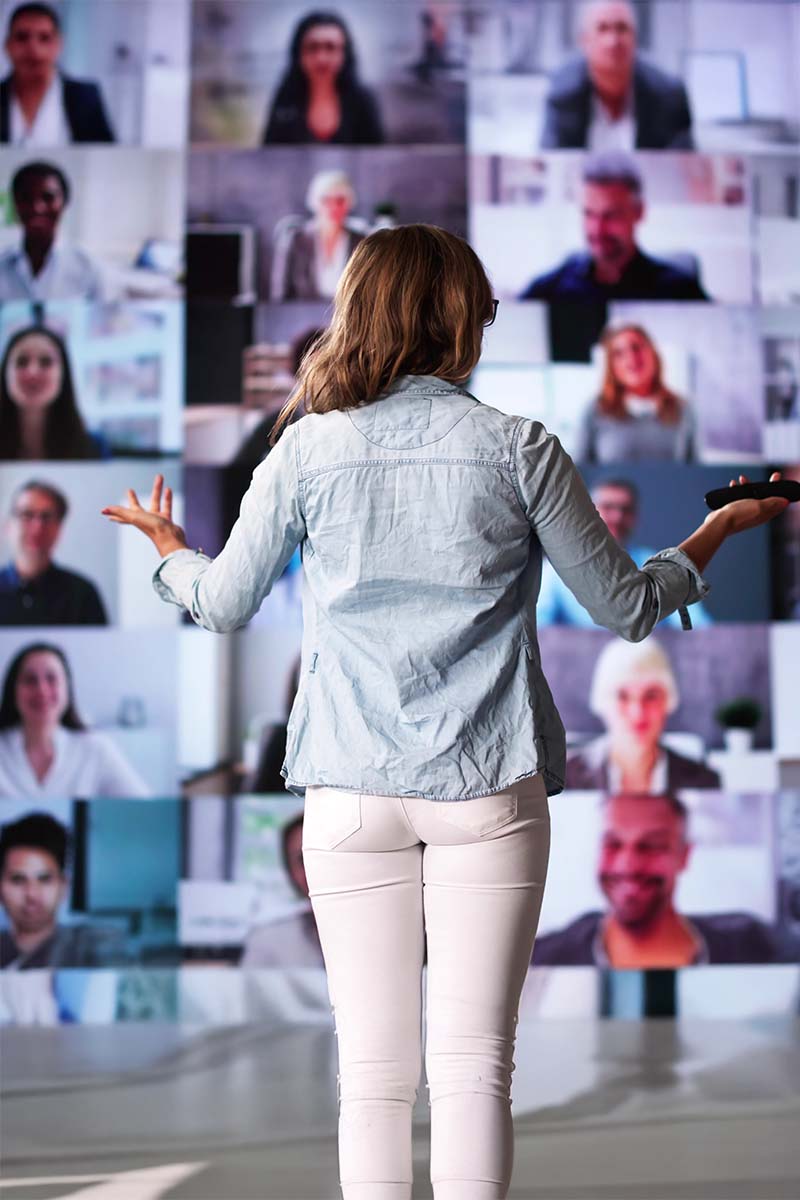 Live event header 800x1200px Lady standing presenting in front of a mulitiple screen of attendess