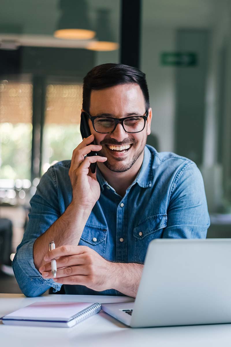 Momentum Min Admin 800x1200px Bearded male on mobile in front of laptop with smiling and happy with the conversation he is having