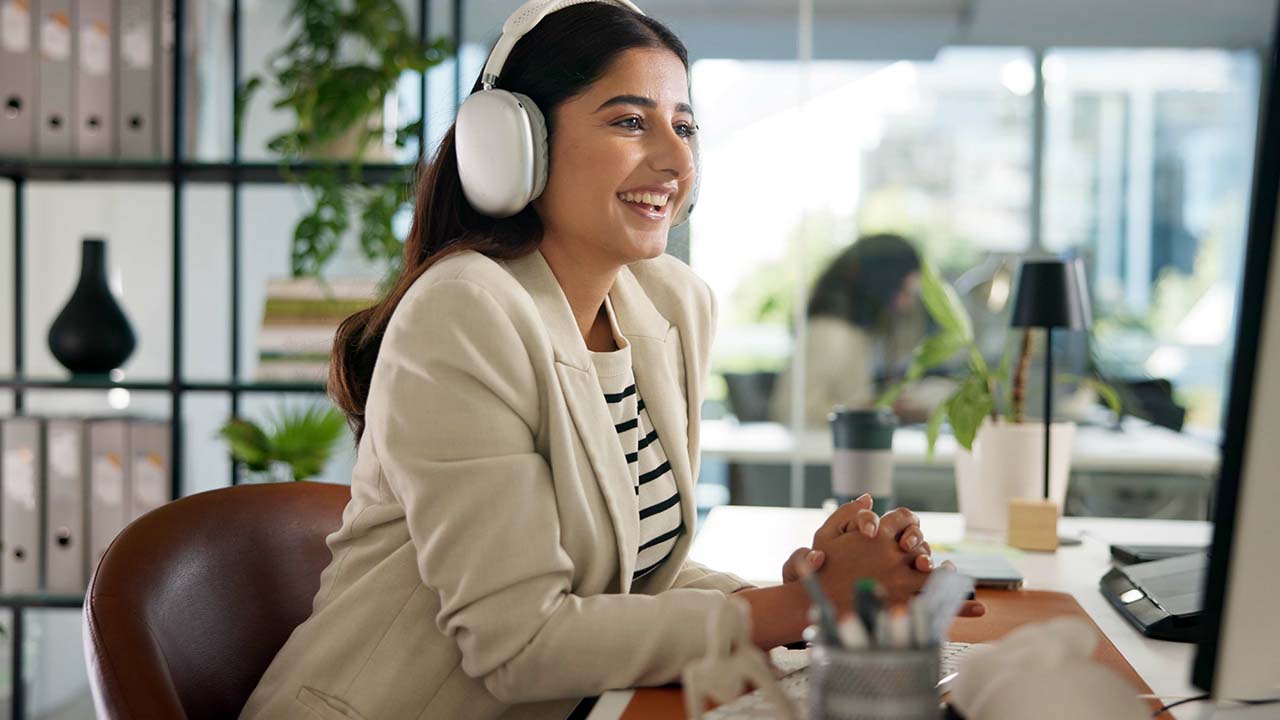 Onboarding 1280x720px Young professional female dressed in corporate attire with headphones on at desk watching and listening to an online employee induction event