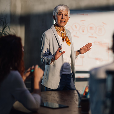 Resilence & agility mobile 400x400px Grey headed lady standing and calmly presenting in front of a whiteboard, looking in control