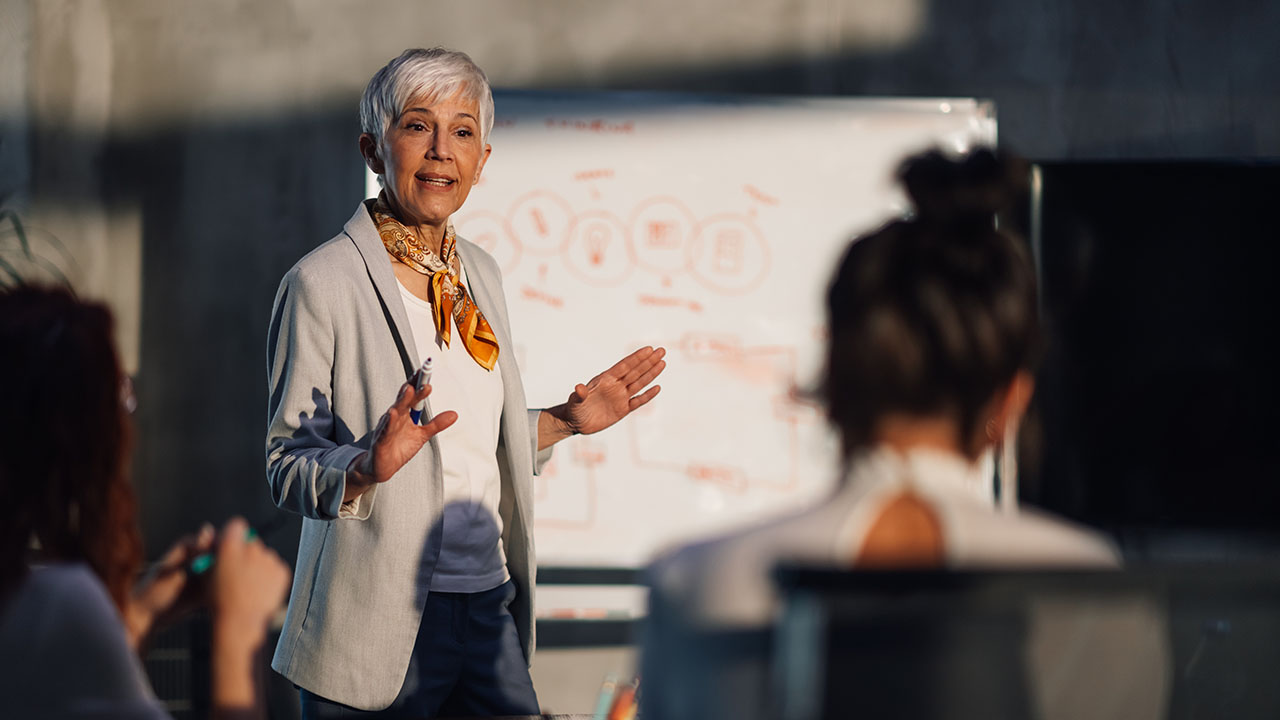 Resilence and agility 1280x720px Grey headed lady standing and calmly presenting in front of a whiteboard, looking in control